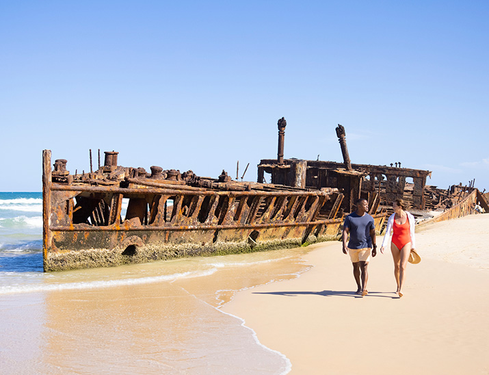 A couple walk along the beach with the Maheno shipwreck behind them